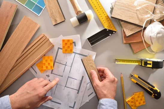 Un homme examine des plans d'architecture sur une table de travail, tenant un &eacute;chantillon de parquet